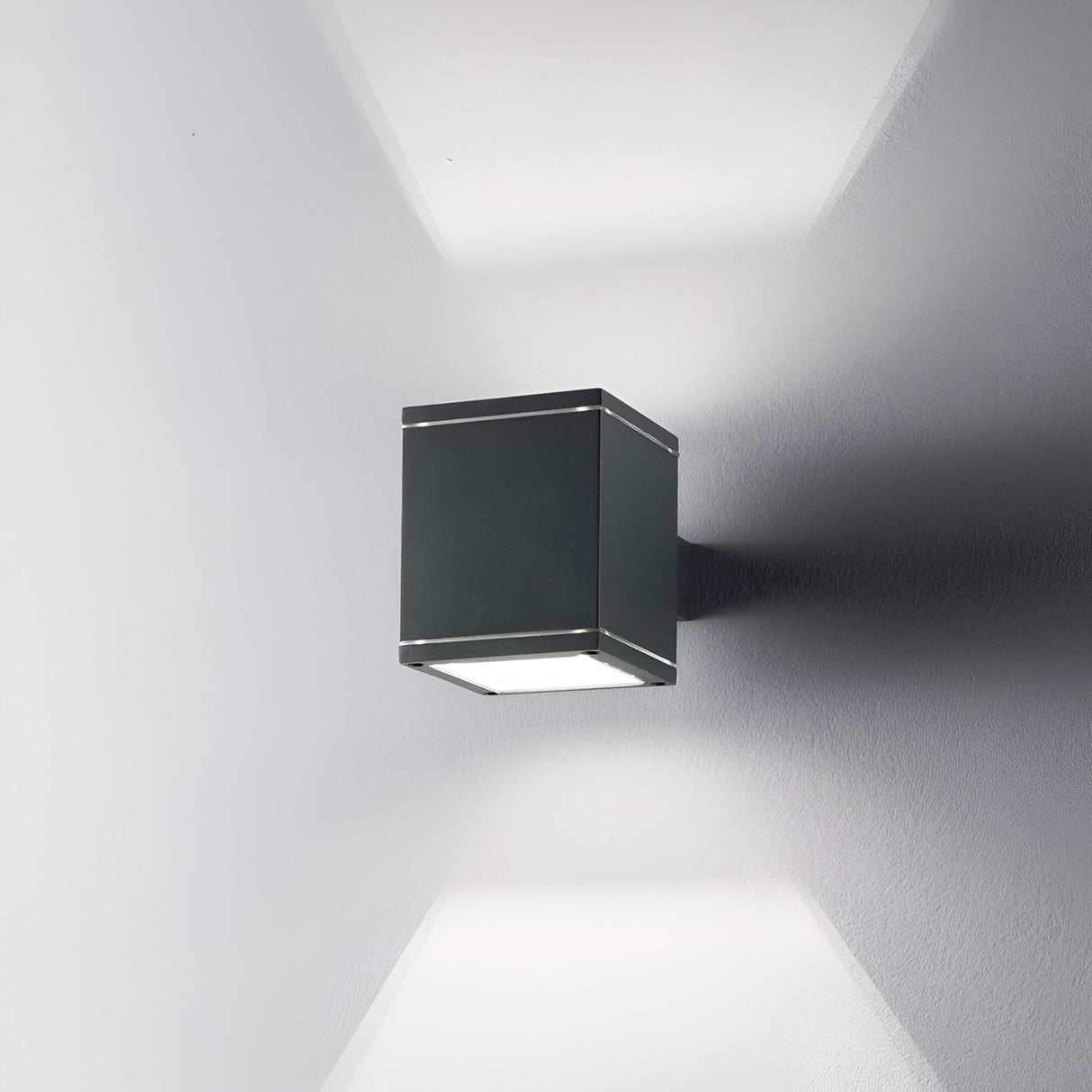 The Opus Square Outdoor Wall Light in anthracite, with its modern cube shape, is mounted on a white wall. It casts light both upward and downward through Pyrex glass, creating a geometric pattern of light and shadows.