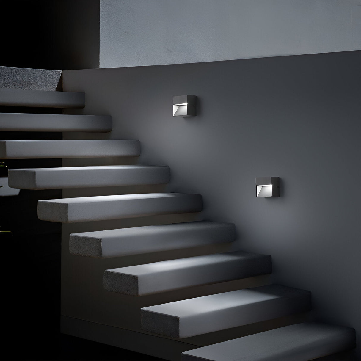 A dimly lit, modern staircase features sleek Jupiter-2 Outdoor Wall Lights - Grey on a dark gray wall. The rectangular steps seem to float, enhancing the minimalist and contemporary style. These weather-resistant lights contribute durability to the chic ambiance.
