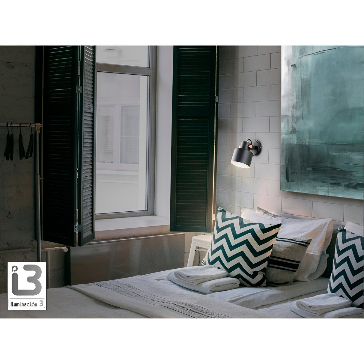 A cozy bedroom with a neatly made bed, chevron-patterned pillows, large windows with dark shutters, and natural light. The Erika Single Spotlight in Matte Black/Satin Copper is mounted on the wall. A painting and the Iluminación 3 logo complete the scene.