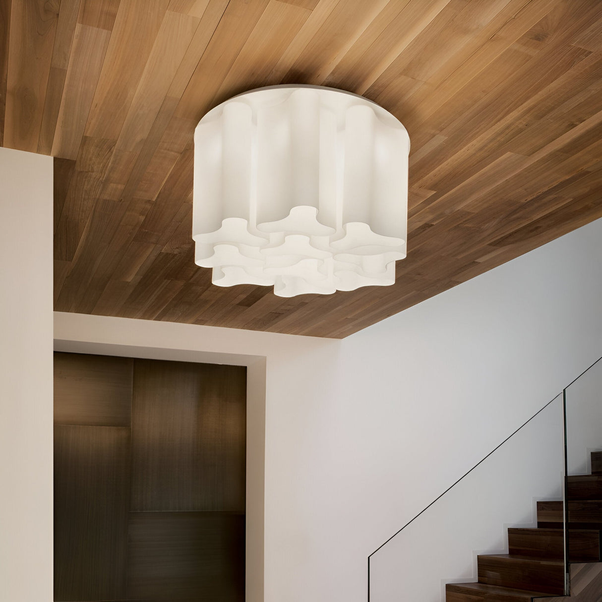 The Haze 10 Light Glass Flush in white, with its wavy, sculptural design, is mounted on a wooden ceiling and features elegant glass elements. The white fixture contrasts beautifully with the dark wood panels. A staircase with a glass railing is partially visible on the right, enhancing the sleek aesthetic.
