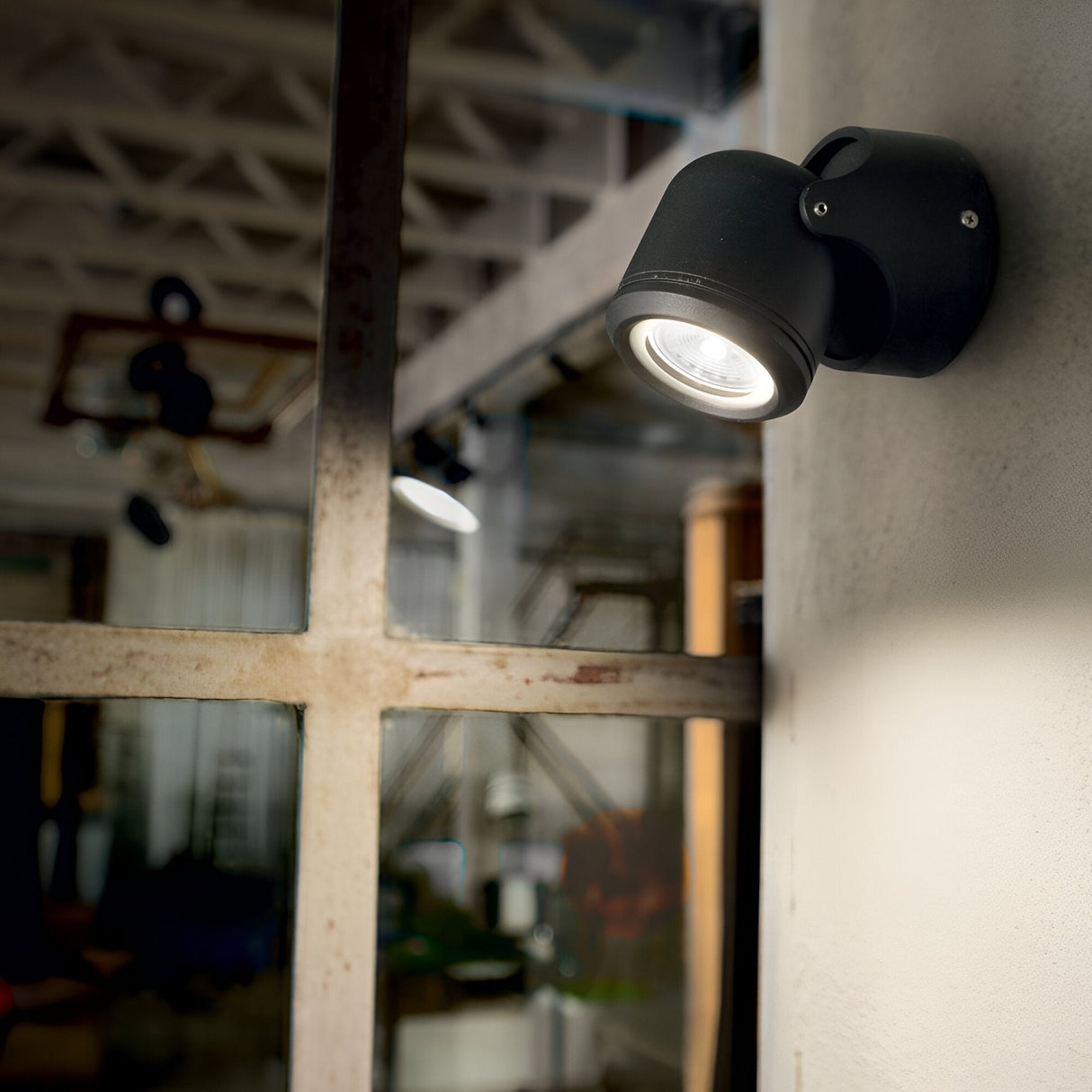 The Vista Outdoor Wall Spotlight - Black is illuminated next to a window, casting light on the rustic interior with exposed beams and glass. This wall-mounted spotlight enhances the modern industrial design and boasts an IP44 rating for durability in varied conditions.