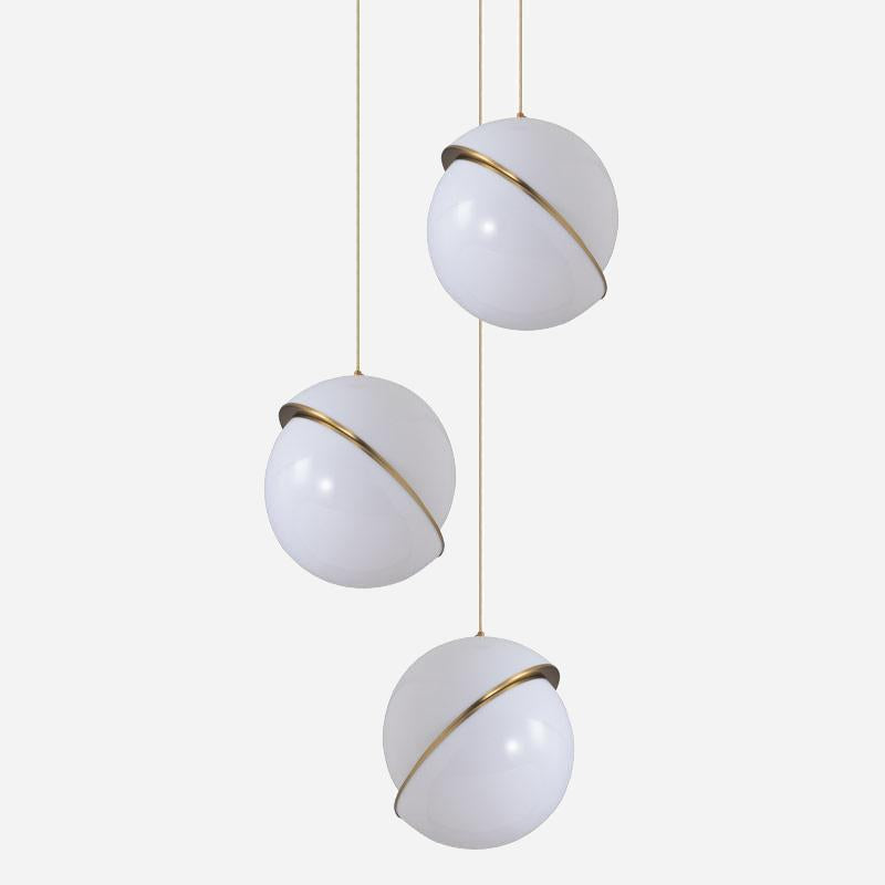 The Arc Pendant Light - Gold & White features three white globes suspended at varying heights. Each globe is elegantly encircled by a thin brushed brass band, offering a sleek and contemporary look against a plain light gray background.