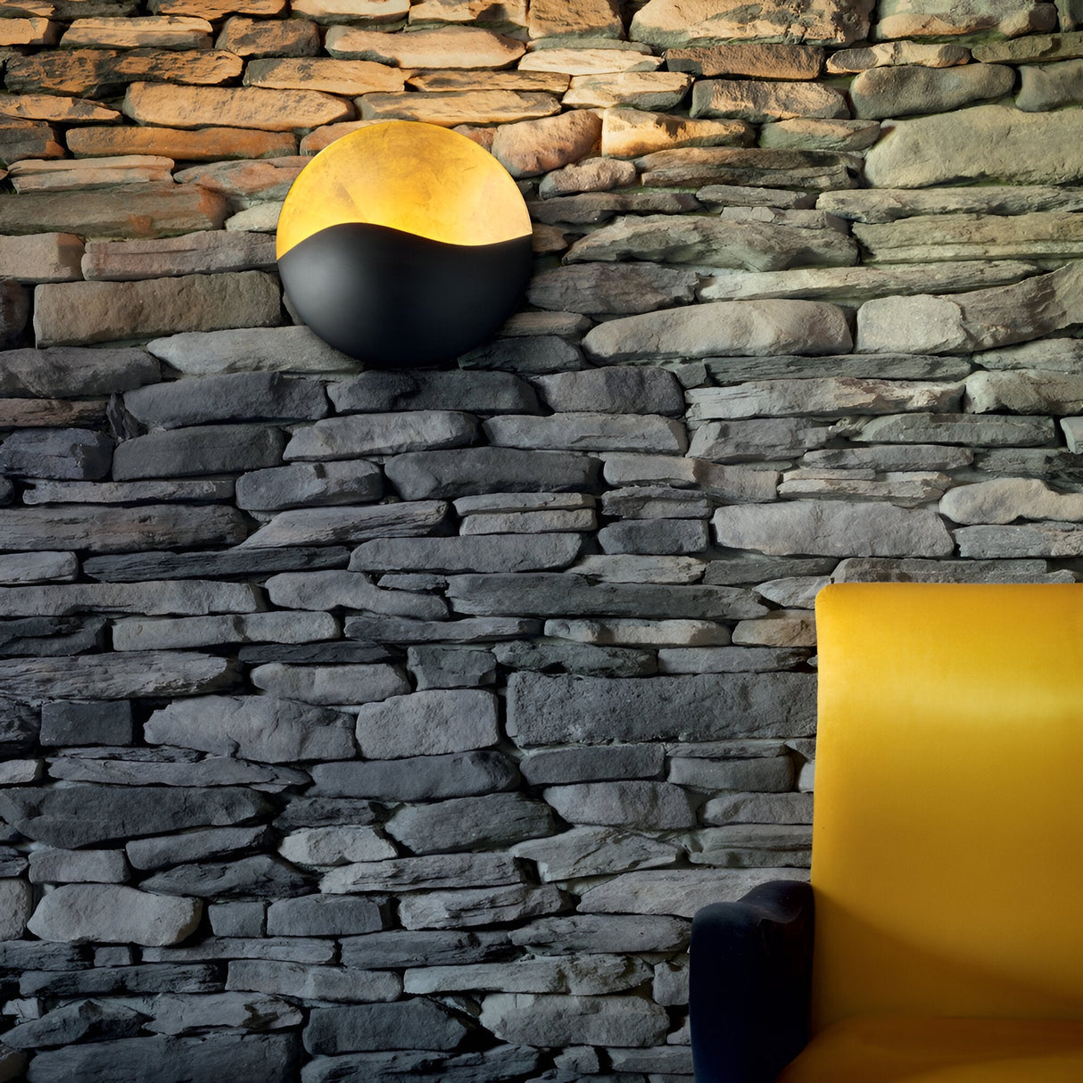 The textured stone wall features a Beamrise 4 Light Wall Light in gold, casting a warm glow. Below, the edge of a modern chair with vivid yellow upholstery adds a pop of color against the rustic stone backdrop. This scene seamlessly blends natural materials with contemporary design elements, accentuated by dramatic ambient lighting.