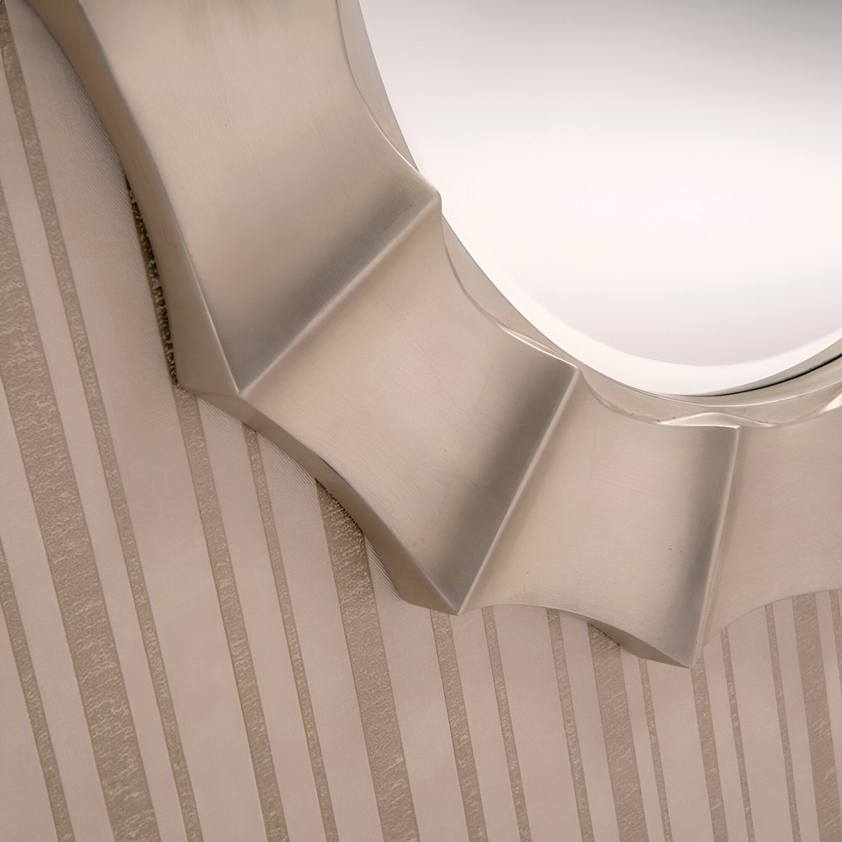 Close-up of the Zenith Circle Wall Mirror Champagne - 104cm, showcasing its metallic frame with a wavy design against a wall adorned with vertical striped wallpaper. This piece boasts a champagne finish and angular, curved edges that impart a modern and artistic look.