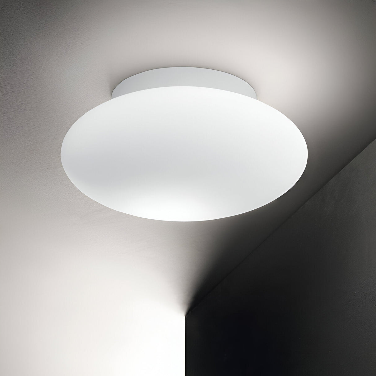 The Serene Outdoor Flush Light - White, with its minimalist circular design, casts a soft glow in the corner of a room adorned with white and gray walls. Its sleek, modern aesthetic enhances the space's contemporary vibe while providing durability akin to weather-resistant outdoor flush lights.