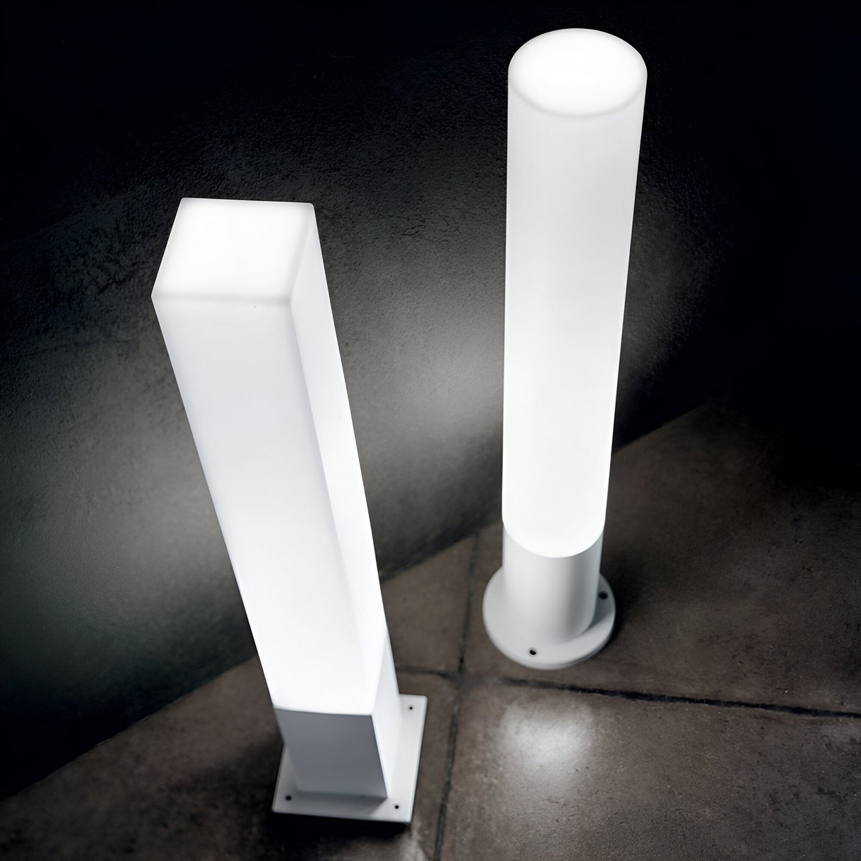 Two freestanding lamps with modern designs, including the Lumira Round Outdoor Bollard Light - White with its durable cast aluminium frame, are placed on a tiled floor. The square and cylindrical shapes cast a gentle white glow against a dark wall, making them ideal for chic outdoor illumination.