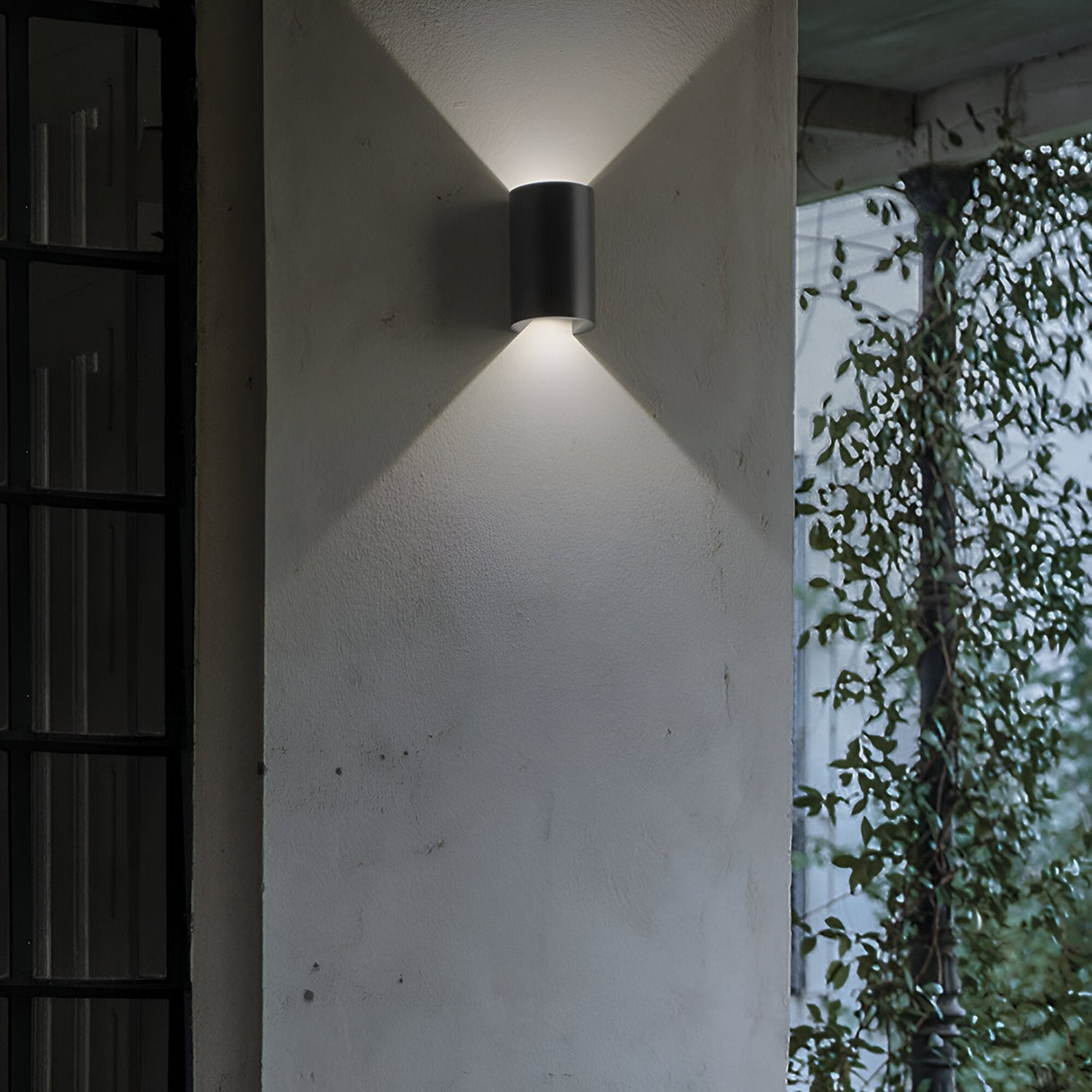 The Peak LED Outdoor Wall Light 6W 4000K - Black, installed on a white exterior wall, delivers a dual light emission that casts a gentle, symmetrical glow both upward and downward. Made from die-cast aluminium, it seamlessly blends into the setting where climbing plants introduce a hint of greenery on the right.