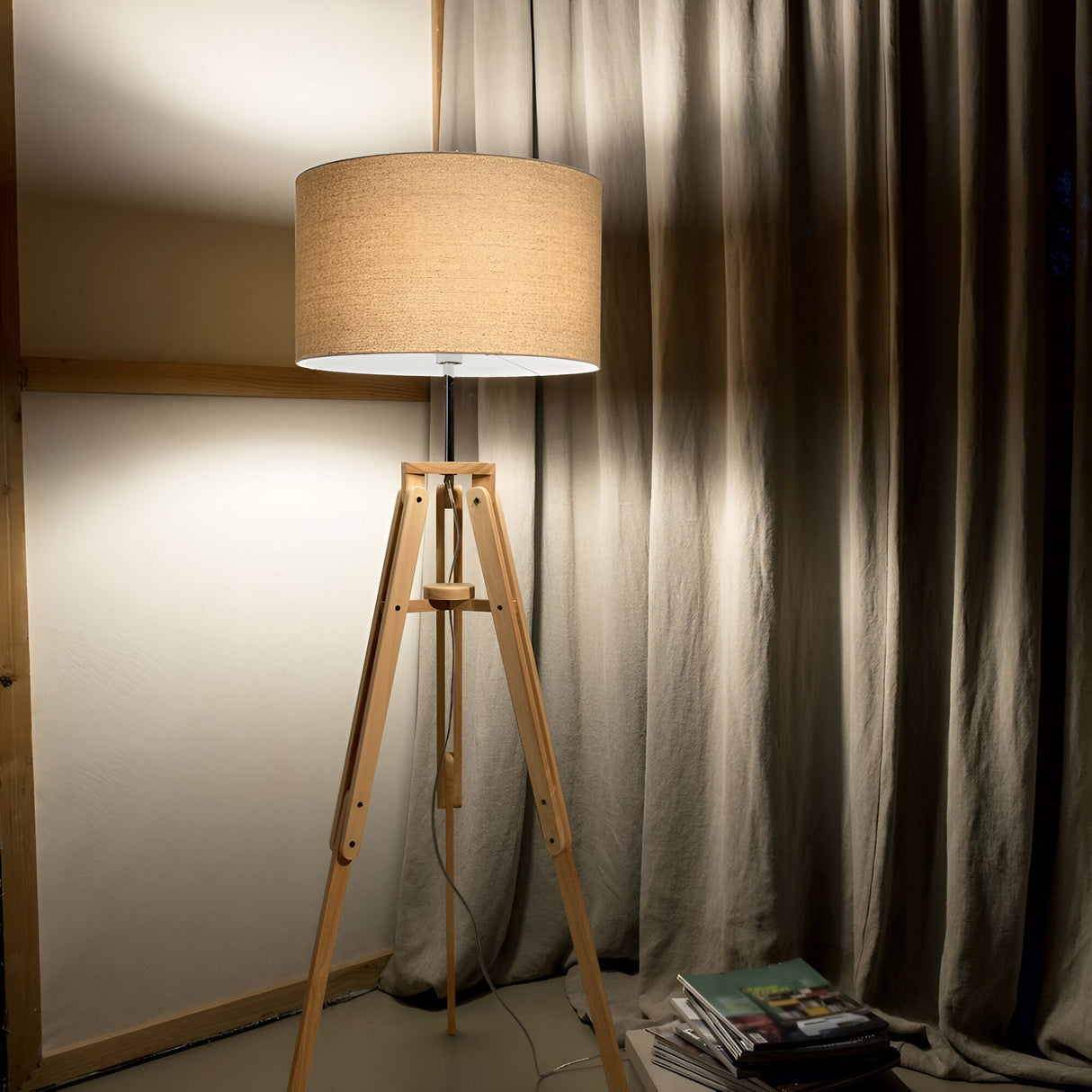 The Gaia Wood Floor Lamp - Light Brown, exuding rustic charm with its natural wood tripod, stands elegantly in a dimly lit room. Behind it, gray curtains drape over the window. A stack of magazines is neatly arranged on the floor to the right of the lamp.