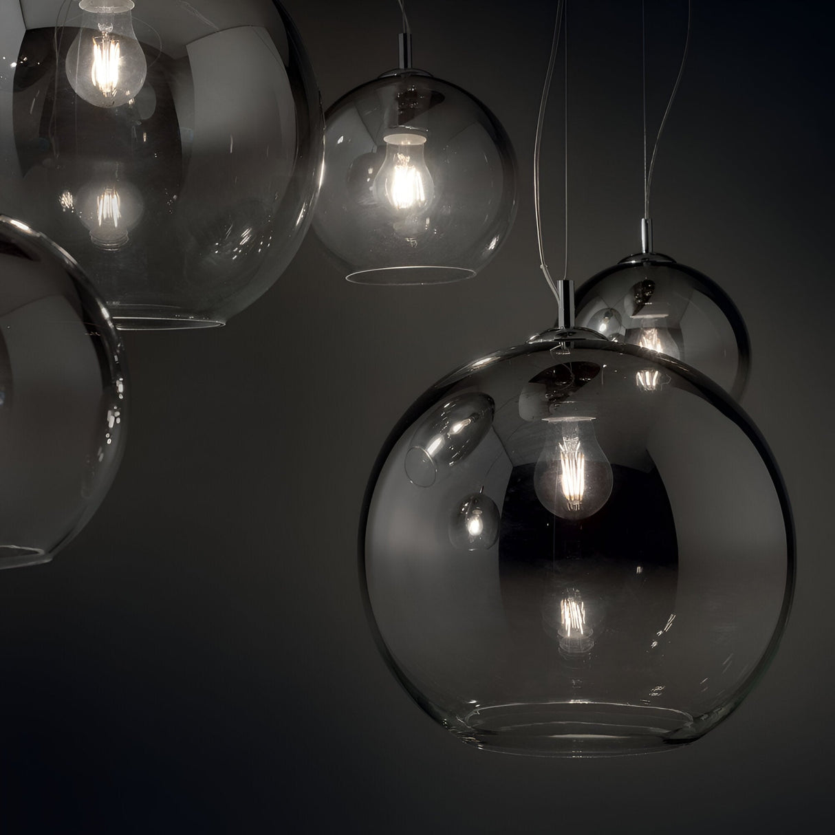 The Emberlyte 5 Light Cluster Pendant - Chrome features glass pendant lights with exposed bulbs against a dark background, casting a warm glow. These modern fixtures have round, transparent designs that create an elegant atmosphere and offer dimmable settings for customizable ambiance.