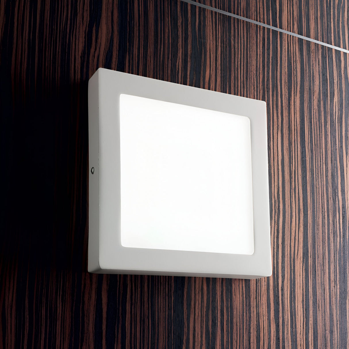 The Zephyrus Square LED Ceiling & Wall Light 3000K in white, measuring 30cm, is mounted on a dark, wood-patterned wall. With its LED bulb turned on, it casts a soft glow across the room.