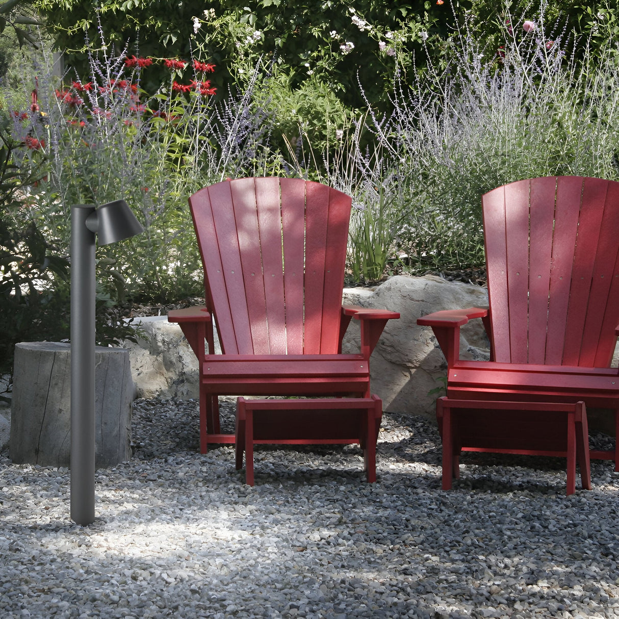 Two red Adirondack chairs with matching footrests sit on a gravel surface, surrounded by lush greenery and lavender plants. The Quasar Outdoor Bollard Light in an anthracite finish stands to the left, while a large rock adds a natural touch to the serene setting.