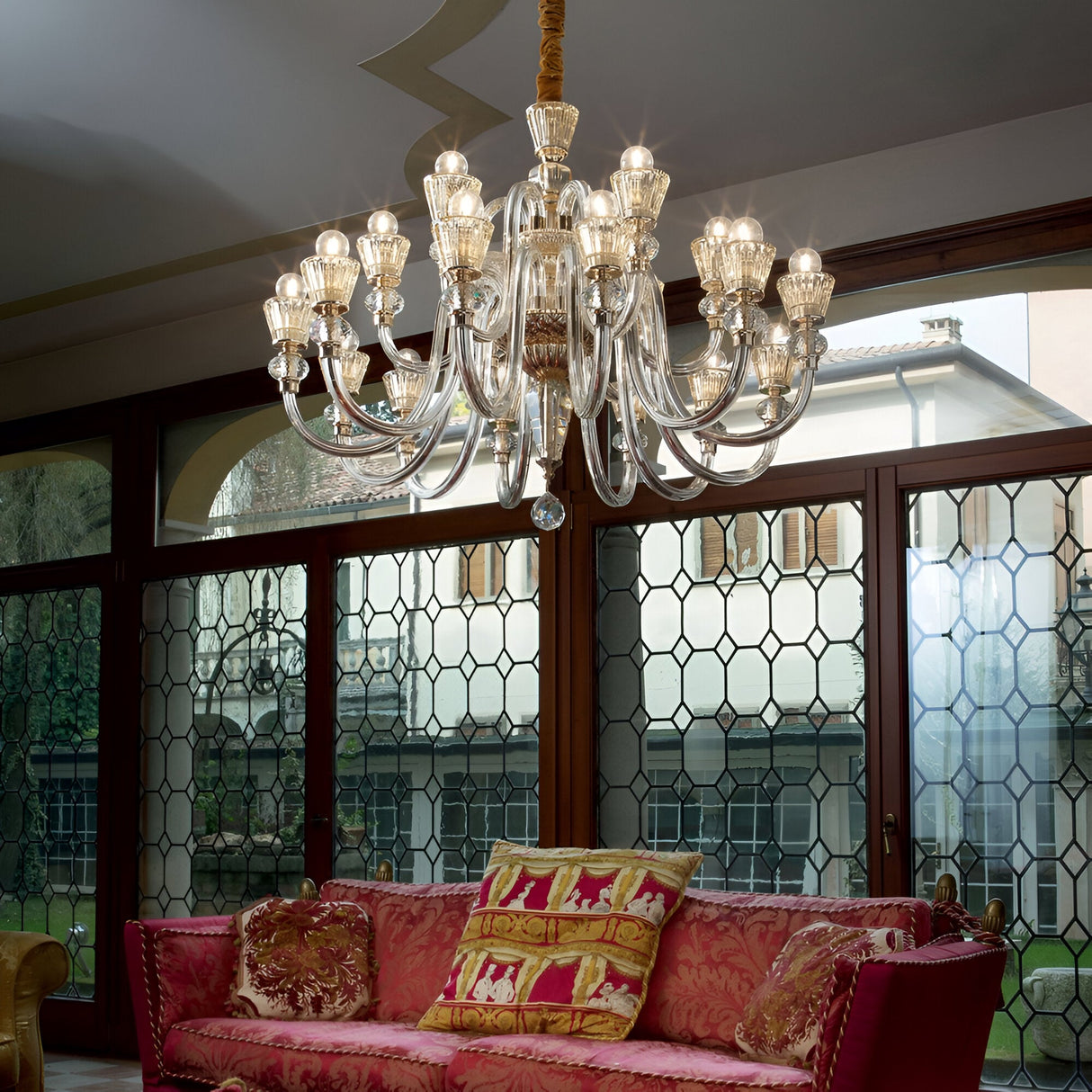 A luxurious, ornate Nexus 18 Light Chandelier in a striking gold and chrome finish, featuring multiple clear glass arms and amber crystal elements, hangs from the ceiling. Below is a richly upholstered red sofa adorned with patterned pillows, set against a backdrop of large windows framed with intricate wooden designs.