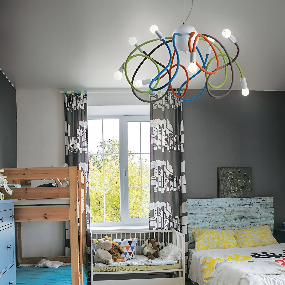 A children's bedroom features a wooden bunk bed on the left and a single bed on the right, decorated with yellow bedding. Overhead, the Radiance 8 Light Suspension in multicolour hangs artfully with its adjustable arms, harmonizing with gray curtains exhibiting a geometric pattern. Stuffed animals are placed on a white bench.