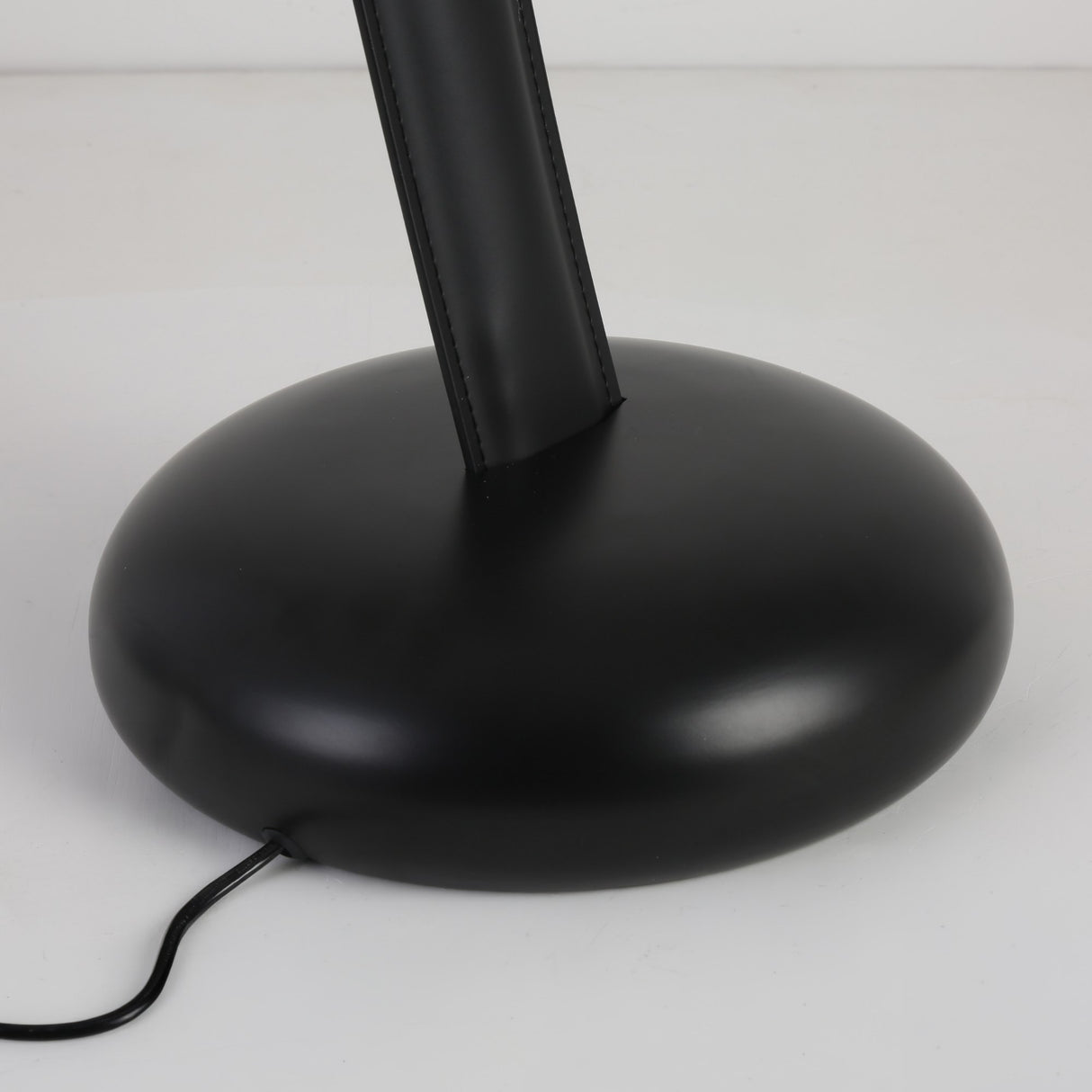 A close-up of the round, black base of the Cordaro 6 Light Leather Floor Lamp - Black, with a black electric cord extending from the bottom across a white surface.