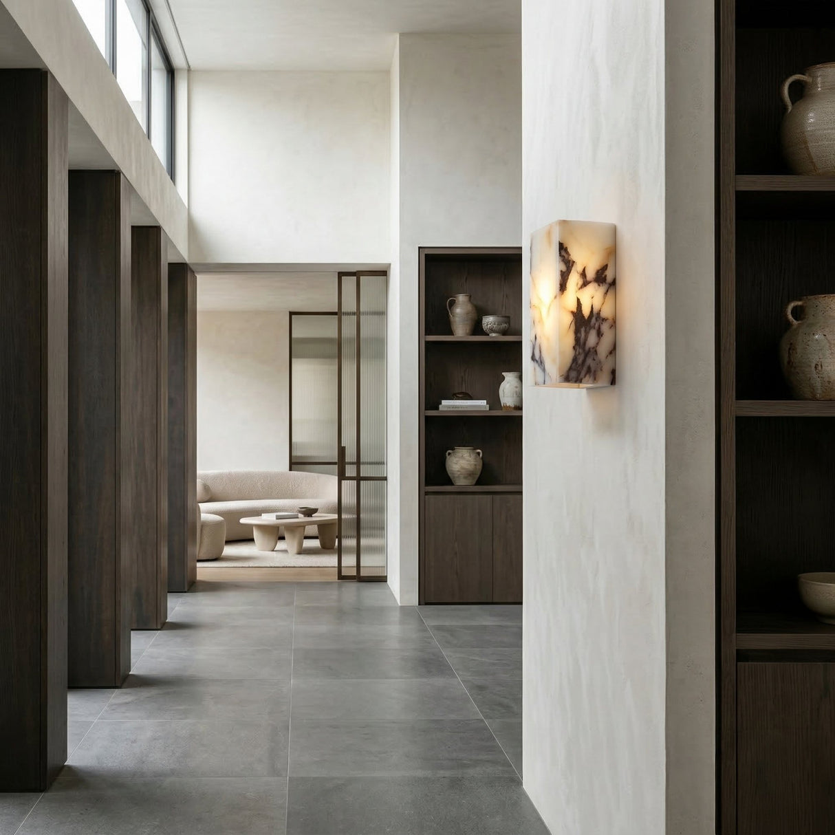 A modern, minimalist interior with gray tile flooring, dark wood shelves holding ceramic vases, the Alvara 1 Light Rectangle Veined Marble Wall Light in white & black, and a glimpse of a beige sofa and round coffee table.