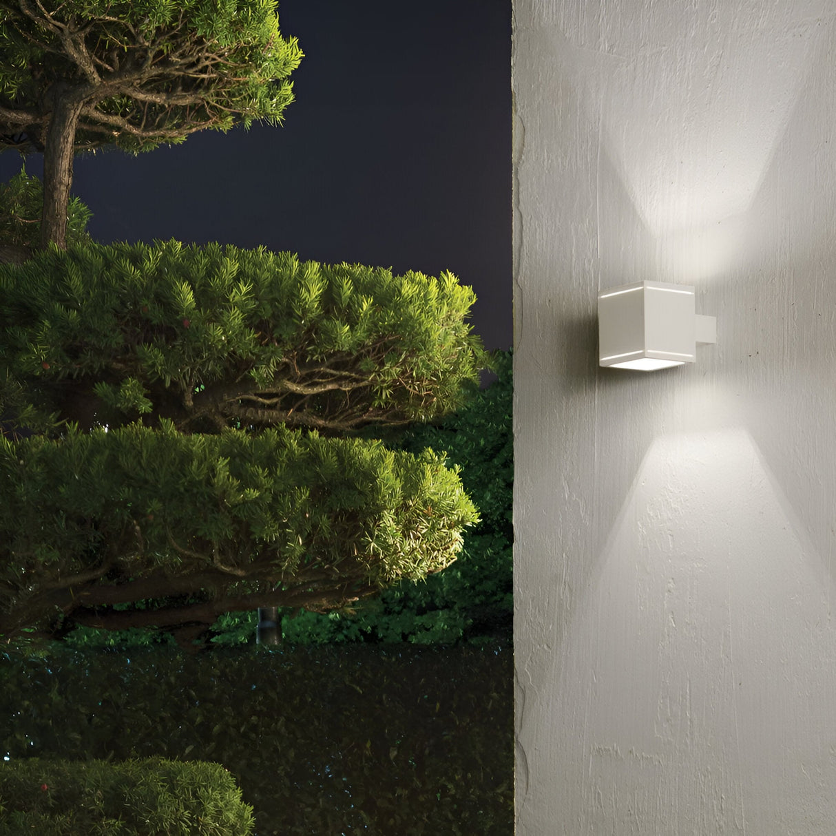 The Opus Square Outdoor Wall Light in white, featuring Pyrex glass, illuminates a textured wall at night and casts a soft glow. To the left, lush green trees with dense foliage stand out against the dark sky, creating a serene outdoor ambiance.