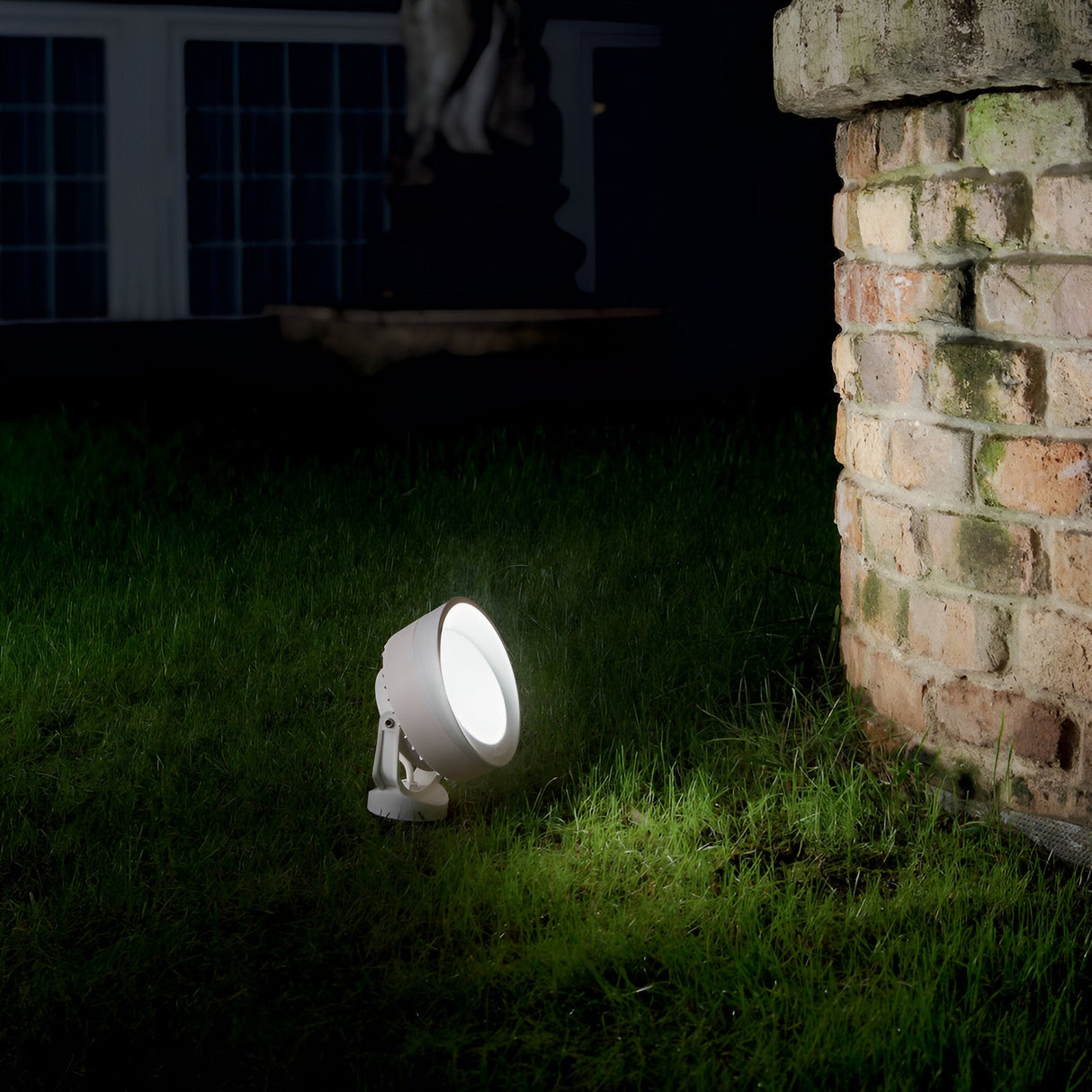 The Point-2 Outdoor Spot Spike Light in White, boasting an IP66 rating, casts light on a patch of grass next to a brick wall at night. In the dark background, windows and a statue are faintly visible, subtly enhanced by the fixture's directional diffuser.