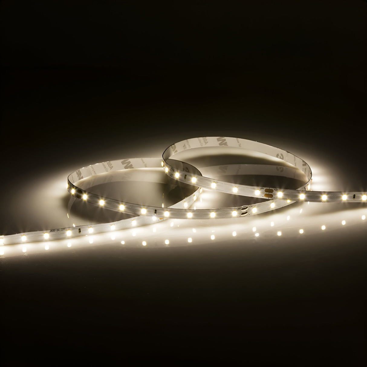 A coiled strip of the 24V LED Strip Light 4000K - 50m softly glows against a dark background. This cool white LED strip is arranged in a loose, overlapping circular pattern, casting a warm and gentle illumination over the area.