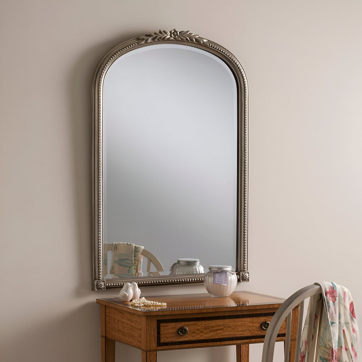 An elegant wooden vanity table with a matching chair is exquisitely adorned with the Mist Arched Wall Mirror in Gold (94x63cm), a delicate jewelry item, and a gold finish candle in a jar. A draped floral cloth complements the scene against the subtly neutral backdrop.