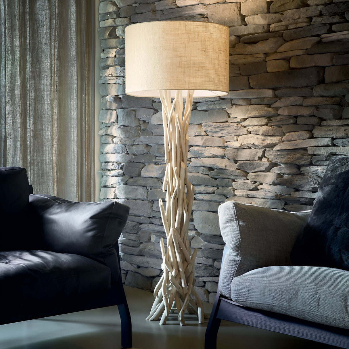 A modern living room showcases the Zone Large Wood Floor Lamp in light brown, featuring a rustic lighting design with a cream shade and an artistically intertwined wooden base. Positioned between two dark sofas against a stone wall, this natural wood lamp creates a cozy and stylish ambiance.