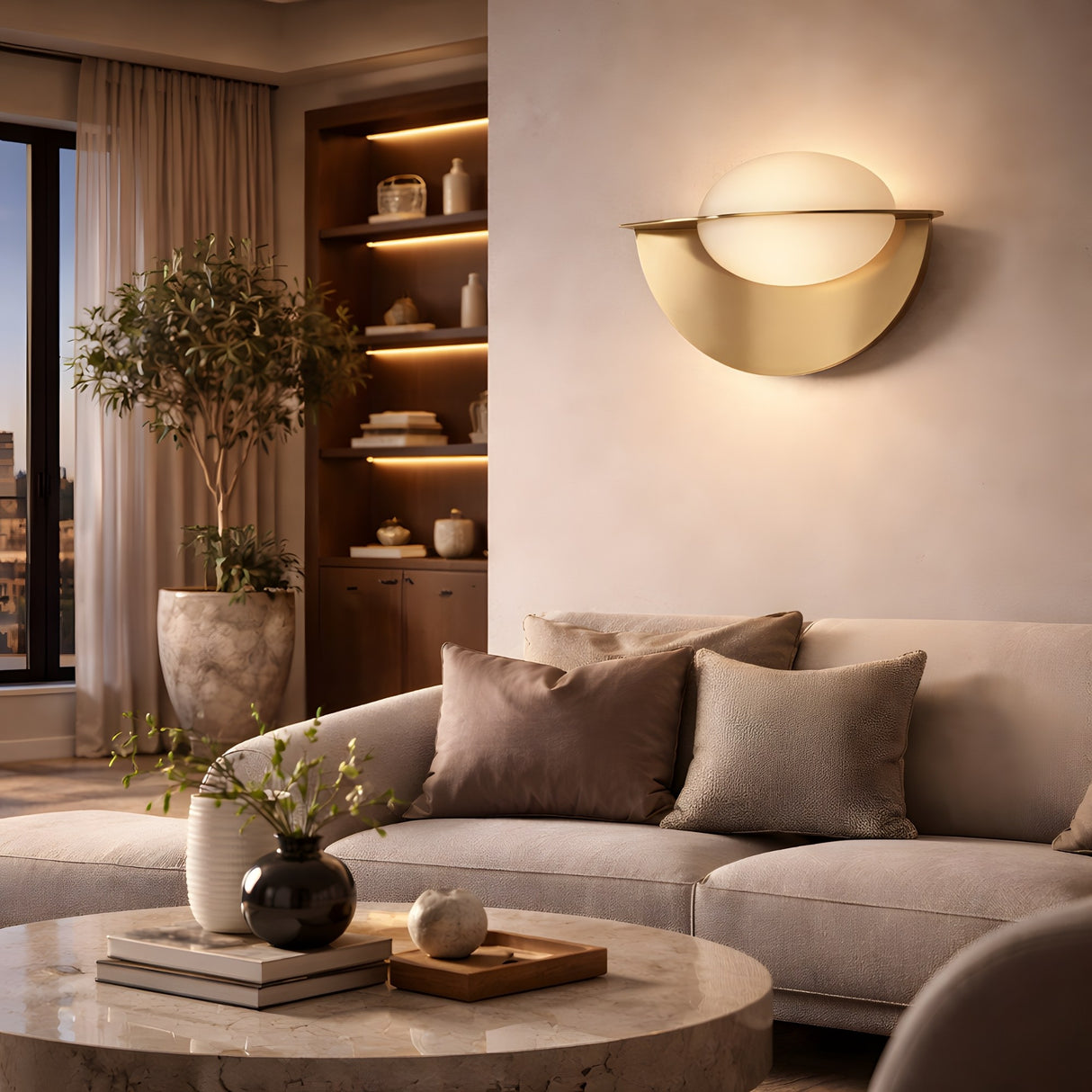 Modern living room with a beige sofa, gray and taupe pillows, a round marble coffee table, decorative vases, potted tree, built-in shelves, and an Althia LED Alabaster Wall Light in brushed brass casting a warm glow on the wall.