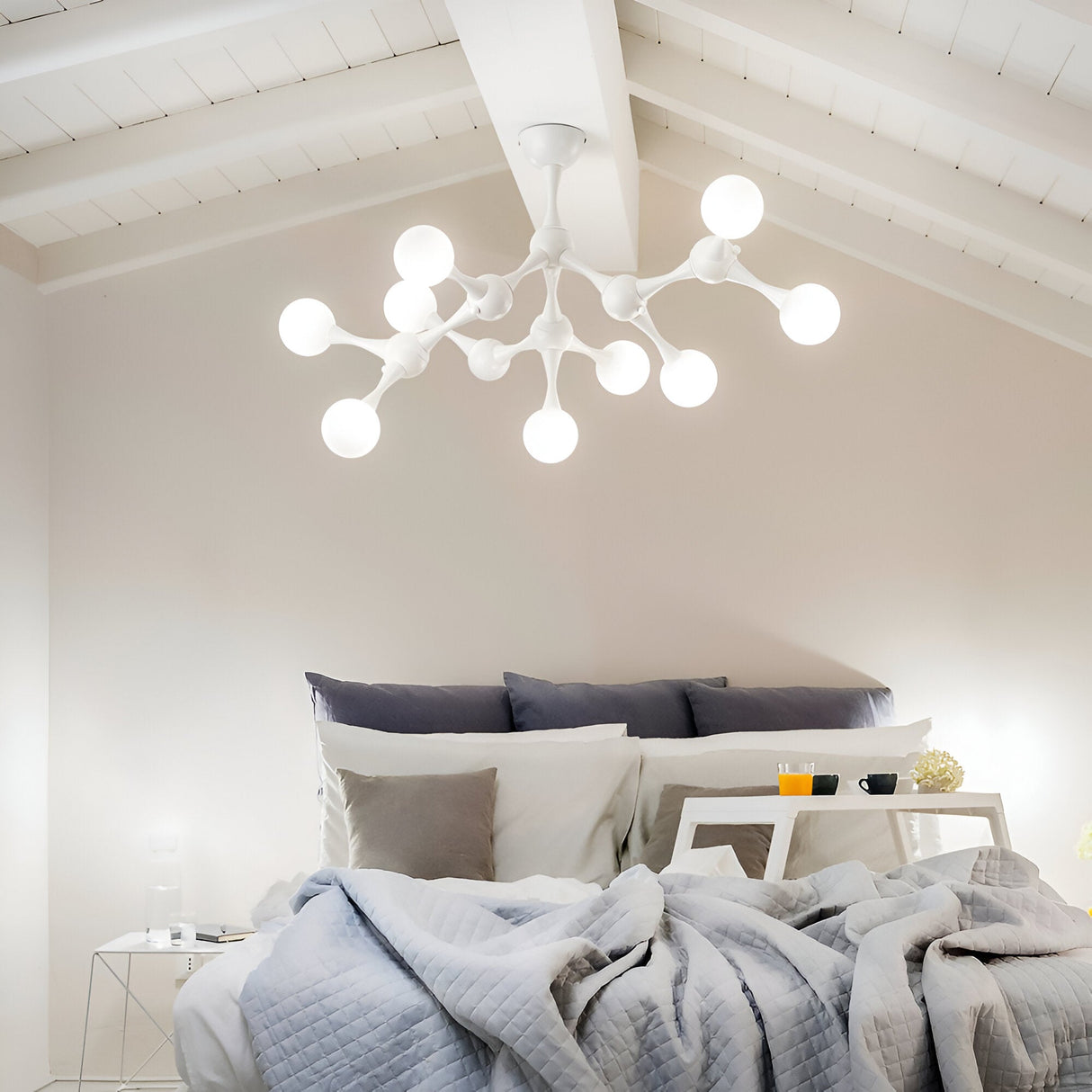 A cozy bedroom is adorned with the Emblazeno 9 Light Semi Flush in white, featuring modern molecular shapes. The bed is neatly made with gray and white bedding, enhanced by dimmable lighting. On a small nightstand sits a lamp and a tray with a glass of orange juice.