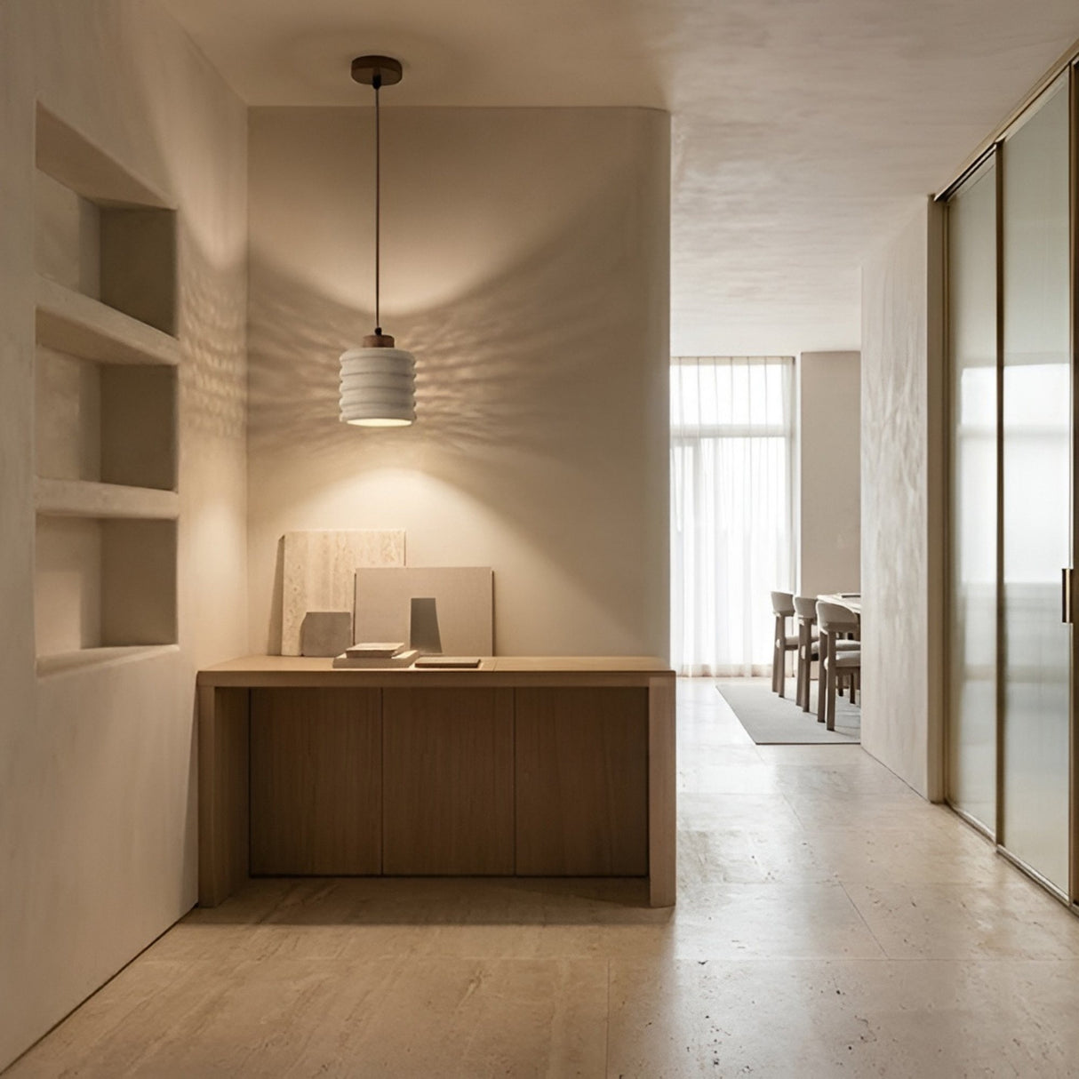 A minimalist, neutral-toned room features a wooden desk, built-in wall shelves, the Rivano 1 Light Ribbed Drum Travertine Single Pendant Light in Walnut Brown & Light Beige, and large frosted glass doors. Sunlight filters through sheer curtains nearby.