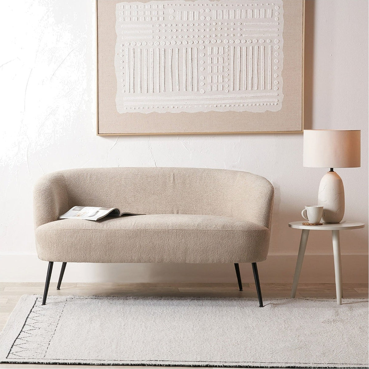 Aviana 130cm sofa in taupe teddy fabric with curved back and black metal legs, styled in a living room beside a side table and lamp.