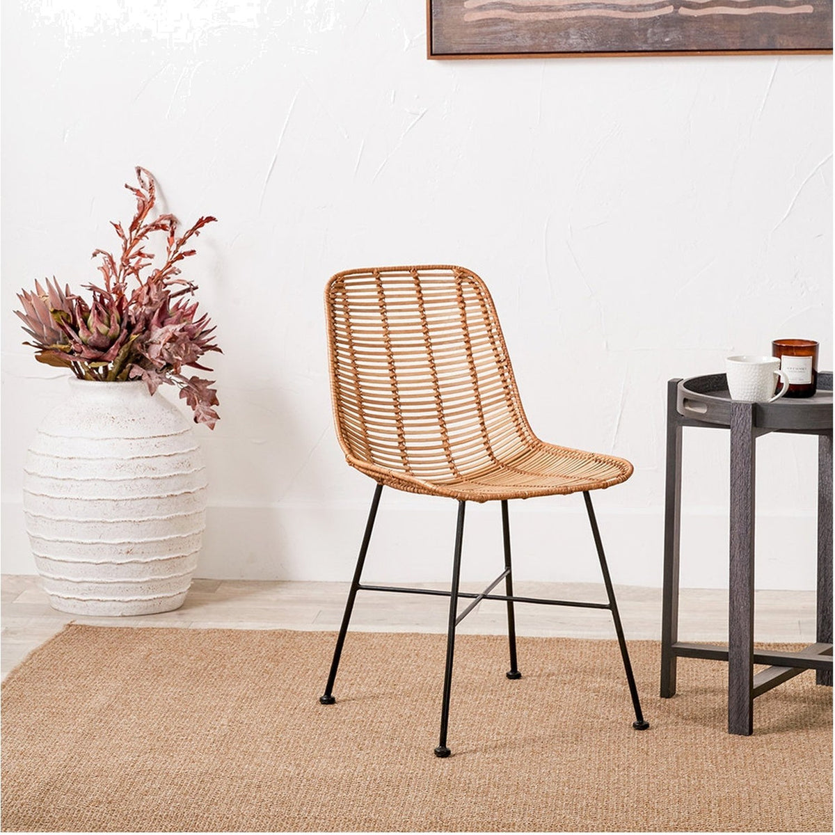 Rattan Core & Black Iron Dining Chair with woven natural rattan seat and back on slim black metal legs, shown in a room setting beside a patterned vase and side table.