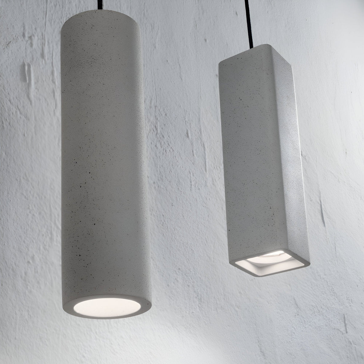 Against a white textured wall, two pendant lights hang: the left one is cylindrical while the right one is the Muse 1 Light Square Concrete Pendant in grey. Both lights embody geometric design principles with their smooth gray materials, casting a soft downward glow that’s ideal for modern interiors.