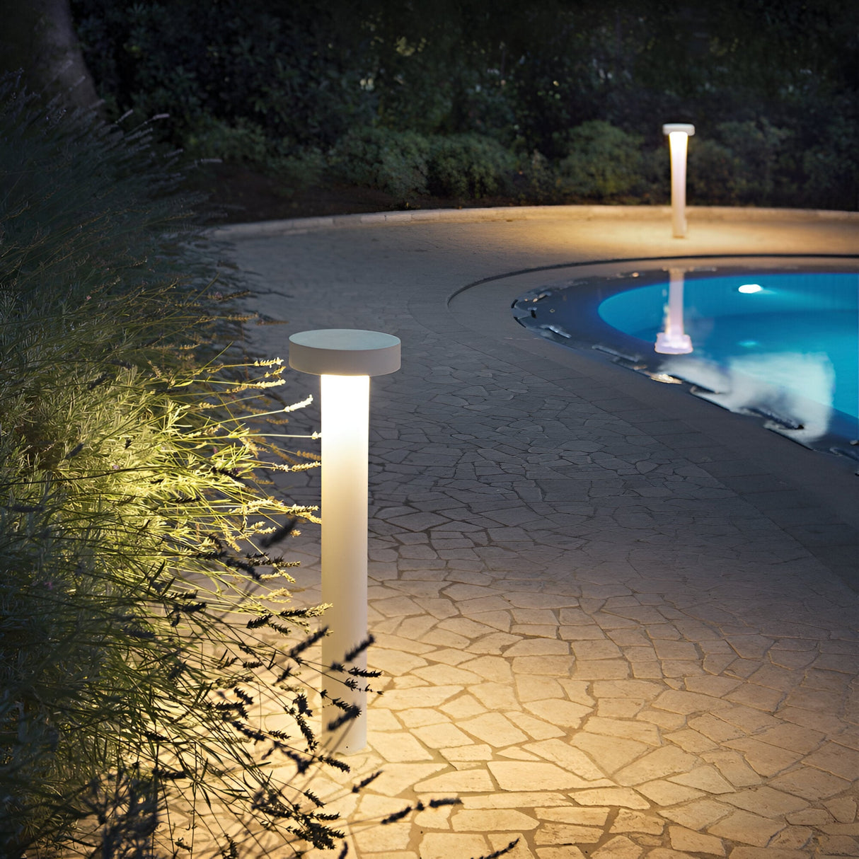 A pathway illuminated by Gloara Outdoor Bollard Light - White, 80cm surrounds a pool at night. The path comprises light-colored stones, flanked by dense, dark foliage. The water in the pool reflects the light, crafting a peaceful ambiance perfect for modern gardens.