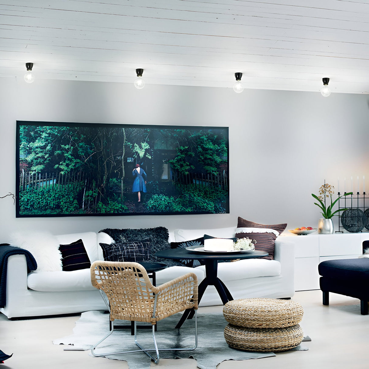 In a modern living room, a sleek black Prismis Ceiling Light with small exposed bulbs adorns the white ceiling. A large artwork depicting a forest scene enhances the gray wall. The space includes a white sofa, a wicker chair, an elegant black enamel round coffee table, and a woven pouf.