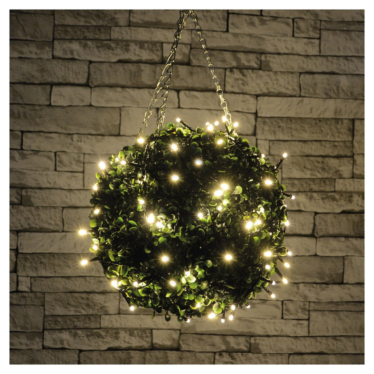The LYYT LED Battery Topiary Ball Light in Warm White hangs in front of a textured stone wall, casting a cozy and decorative ambiance.