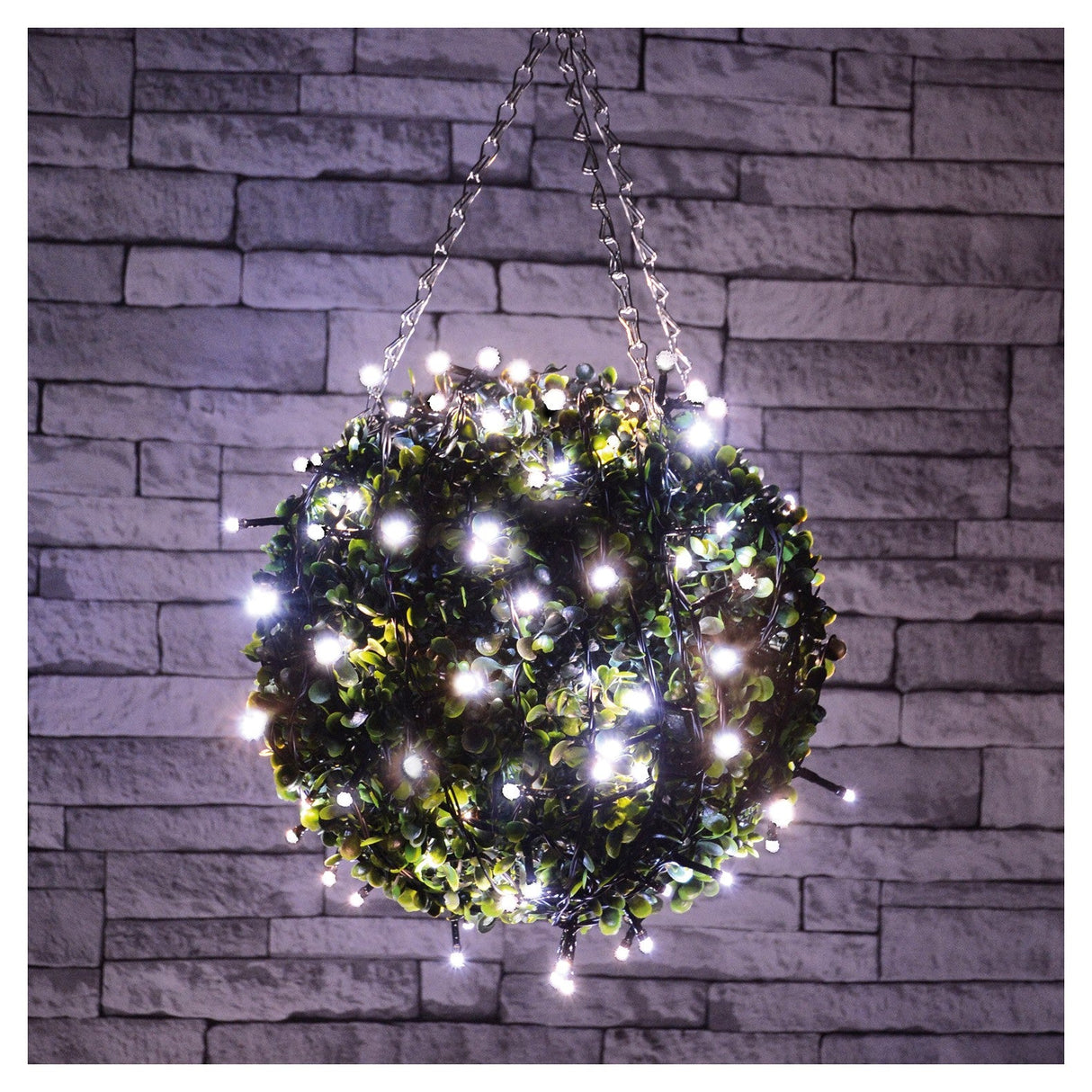 The LYYT LED Battery Topiary Ball Light in Cool White is a spherical decoration crafted from greenery, adorned with numerous energy-efficient LED string lights. Suspended by chains against a textured stone wall, it softly illuminates the space.