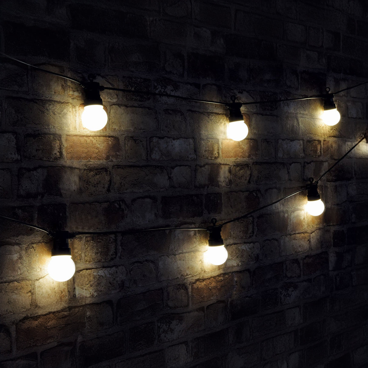 The LYYT Outdoor 10 Festoon Lights, featuring warm white round bulbs, are elegantly draped against a dark brick wall, gently illuminating the scene. These weatherproof LEDs are ideal for outdoor lighting, providing a cozy and welcoming ambiance.