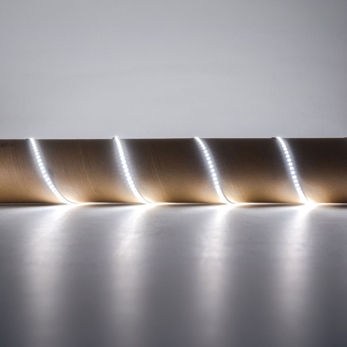 The LYYT 24V High Output LED Strip Light - 6000K emits a bright white light as it coils around a brown cardboard roll, set on a smooth white surface against a plain background. With its self-adhesive backing, this strip guarantees easy application and high luminous flux for superior illumination.
