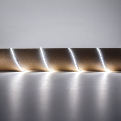 The LYYT 24V High Output LED Strip Light - 6000K emits a bright white light as it coils around a brown cardboard roll, set on a smooth white surface against a plain background. With its self-adhesive backing, this strip guarantees easy application and high luminous flux for superior illumination.