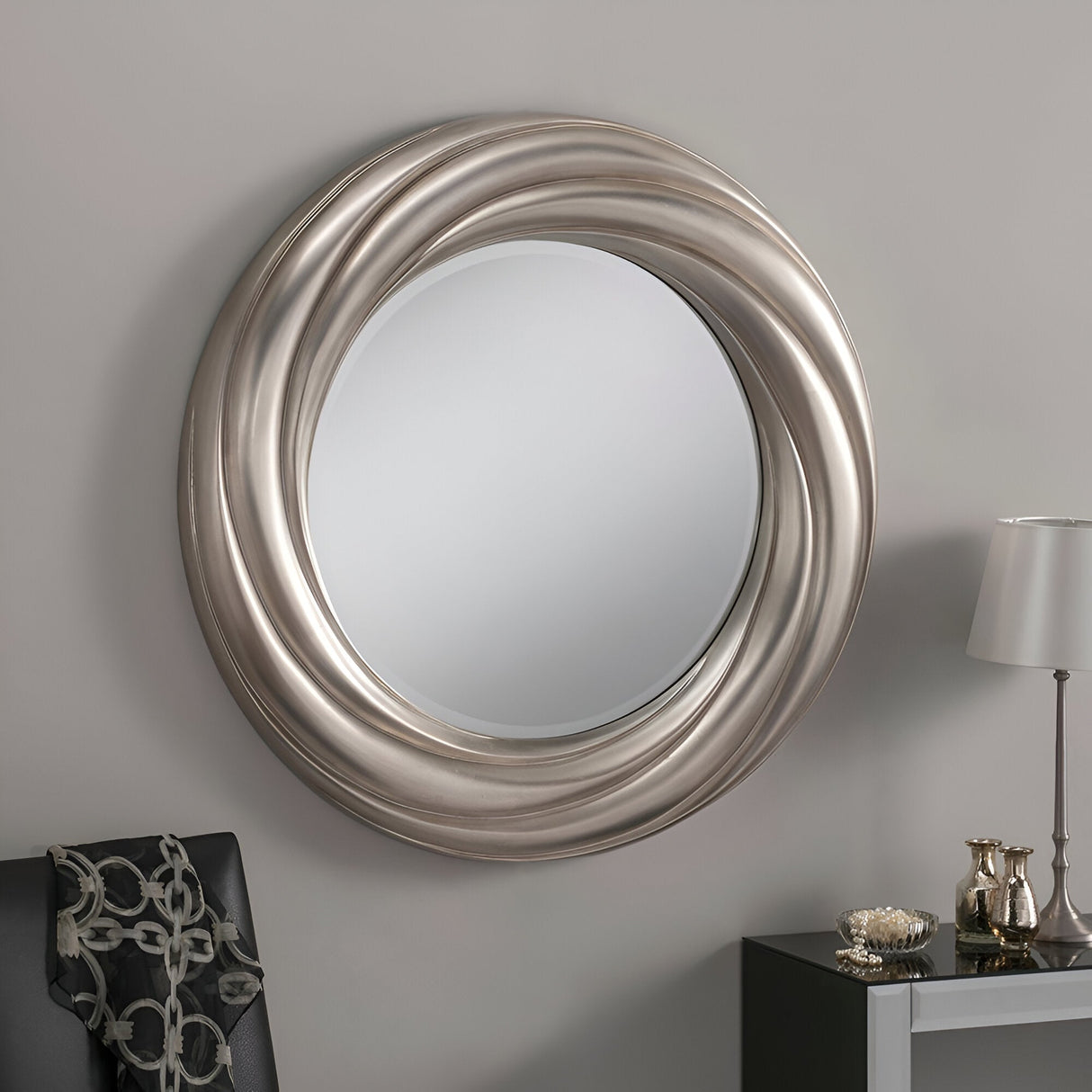 The Iris Circle Wall Mirror Silver - 84cm showcases modern elegance with a chic, metallic silver frame that has a distinctive twisted design. When hung on a light gray wall, it pairs beautifully with the small table beneath it, which is topped with a decorative lamp and various items. The versatile decor is further enhanced by a dark chair draped with a patterned scarf.