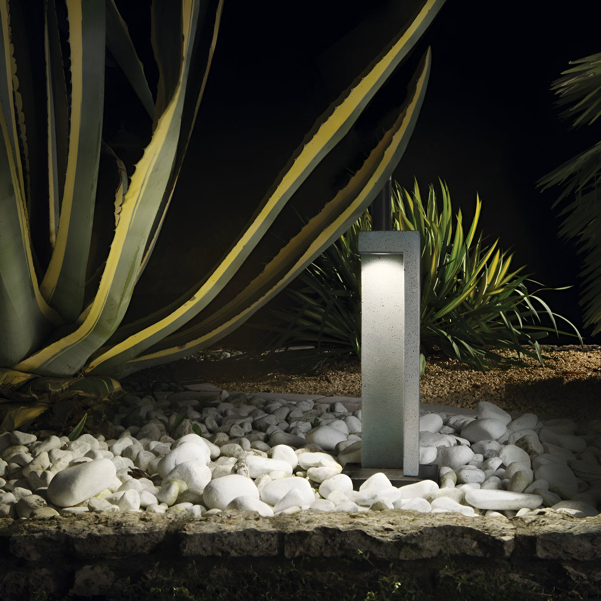 The Starry Outdoor LED Bollard Light 9W 4000K in Grey, standing at 49cm, beautifully illuminates a garden adorned with large, spiky-leaved plants and white decorative stones. Its energy-efficient design enhances the tranquil atmosphere by emphasizing the contrast between the dark foliage and softly lit stone path while ensuring outdoor durability throughout the night.