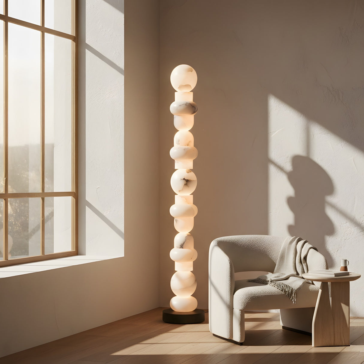 The Stratalux LED Stacked Column Alabaster Floor Lamp in matte black and soft white stands by a cozy white armchair with a blanket, next to a small wooden table in a sunlit minimalist room with large windows and light walls.
