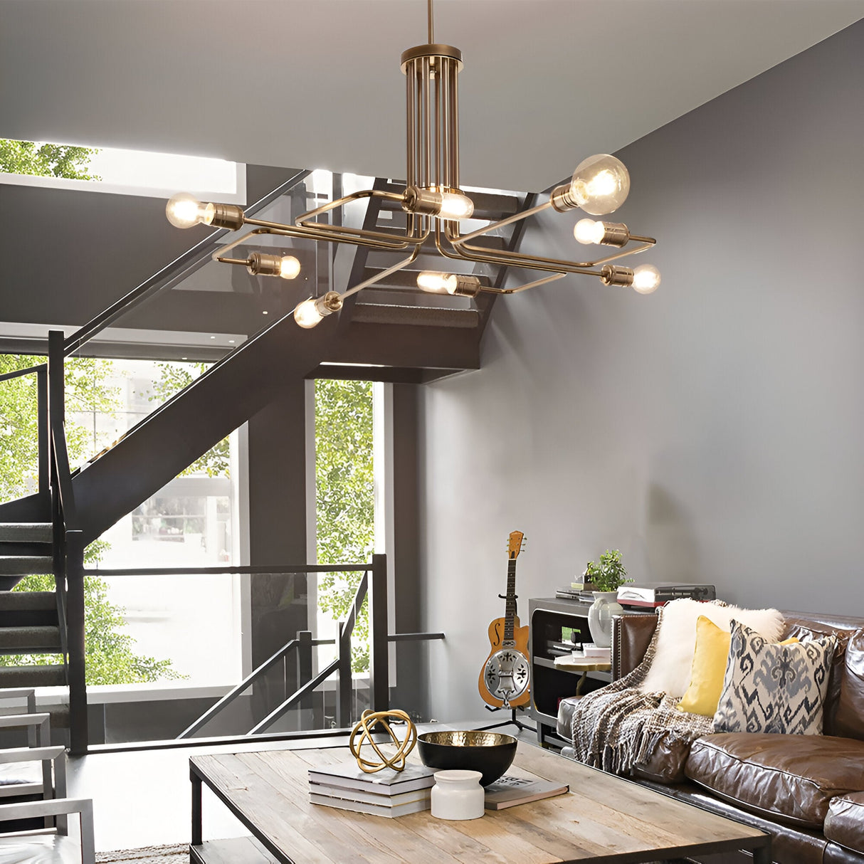 A modern living room showcases the Lustria 8 Light Pendant - Brass, a striking geometric chandelier that uses E27 bulbs and features aged brass accents. The room also includes a brown leather sofa adorned with cushions, a wooden coffee table, bookshelf, and a guitar. A metal staircase provides access to the mezzanine. Expansive windows flood the space with natural light, beautifully highlighting the minimalist decor.