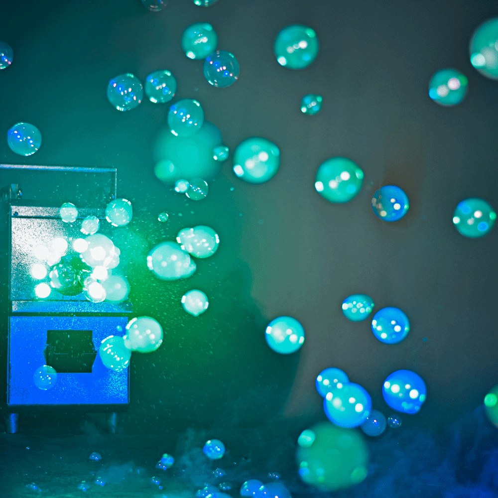 QTX QTFX-LBF2 800W LED bubble and fog machine by AVSL in action, releasing illuminated blue and green bubbles through green-lit fog at an event.