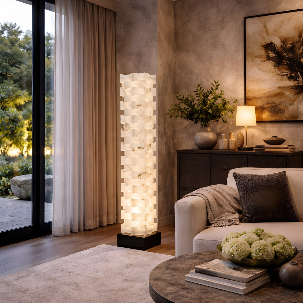A modern living room features the Hexaryn LED Interlocking Alabaster Floor Lamp in Matte Black & Soft White glowing beside a white sofa, with a round coffee table, sideboard decor, and ambient light near large windows with sheer curtains.