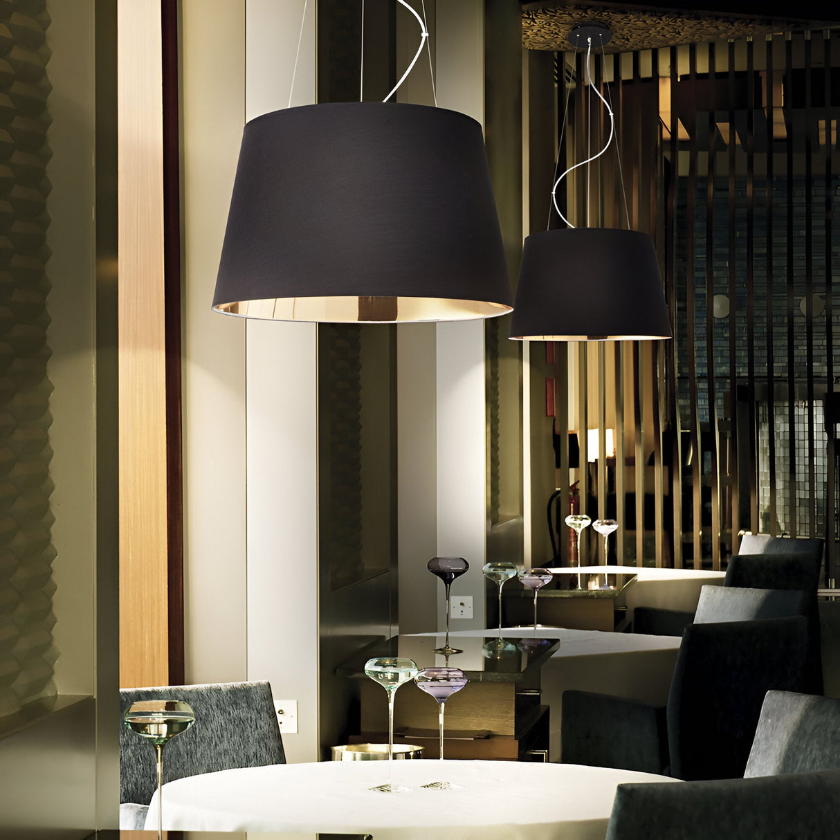 A modern dining area featuring the Hive 6 Light Pendant in black hanging over round tables draped with white tablecloths. The room includes green upholstered chairs and wooden wall accents, providing a contemporary lighting solution that enhances the elegant and cozy atmosphere.