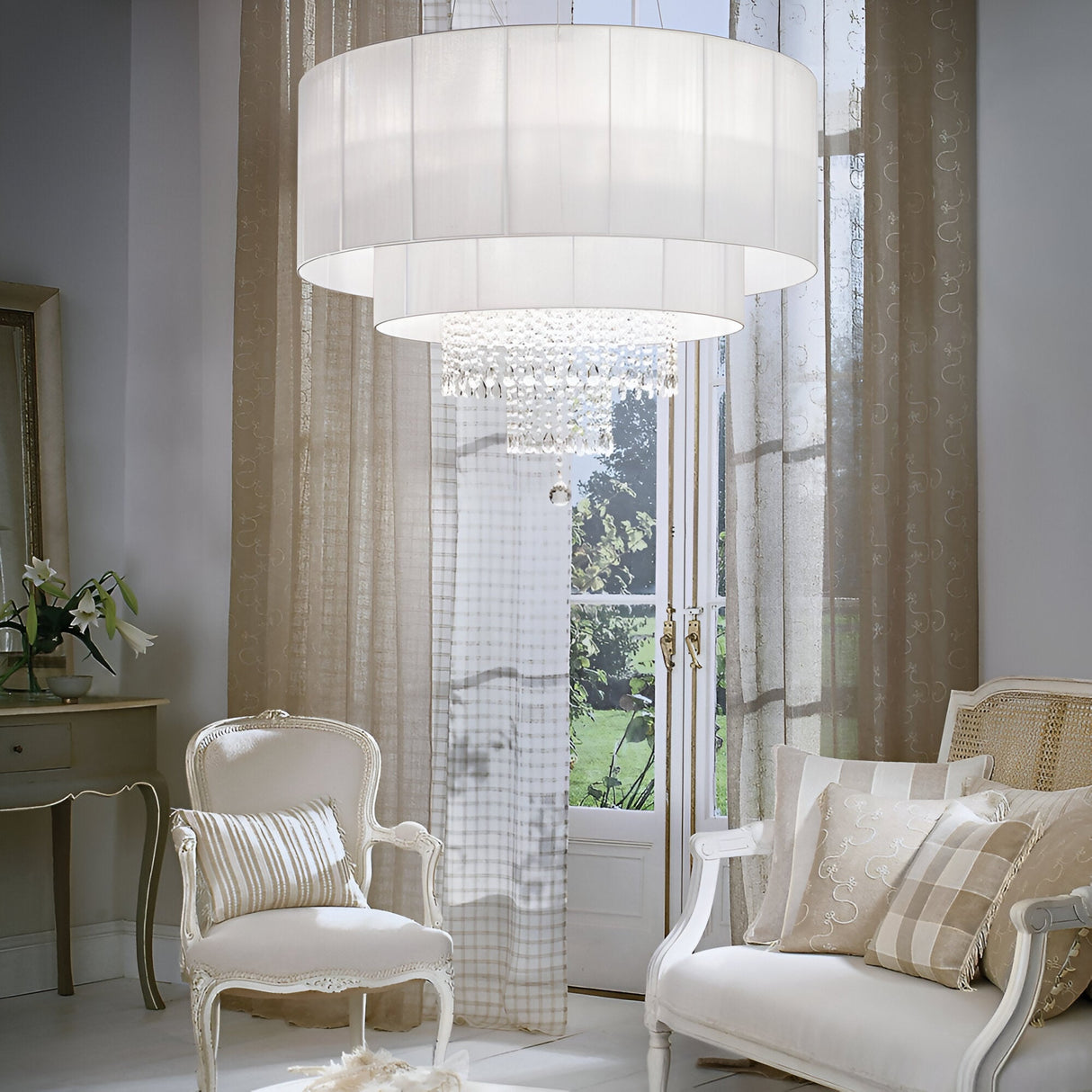 A luxurious living room features the Brillis Crystal 10 Light Chandelier in white, exemplifying elegant lighting with its stunning design. Two ornate white armchairs with patterned cushions are positioned by a window dressed in sheer curtains, inviting natural light into this modern interior.