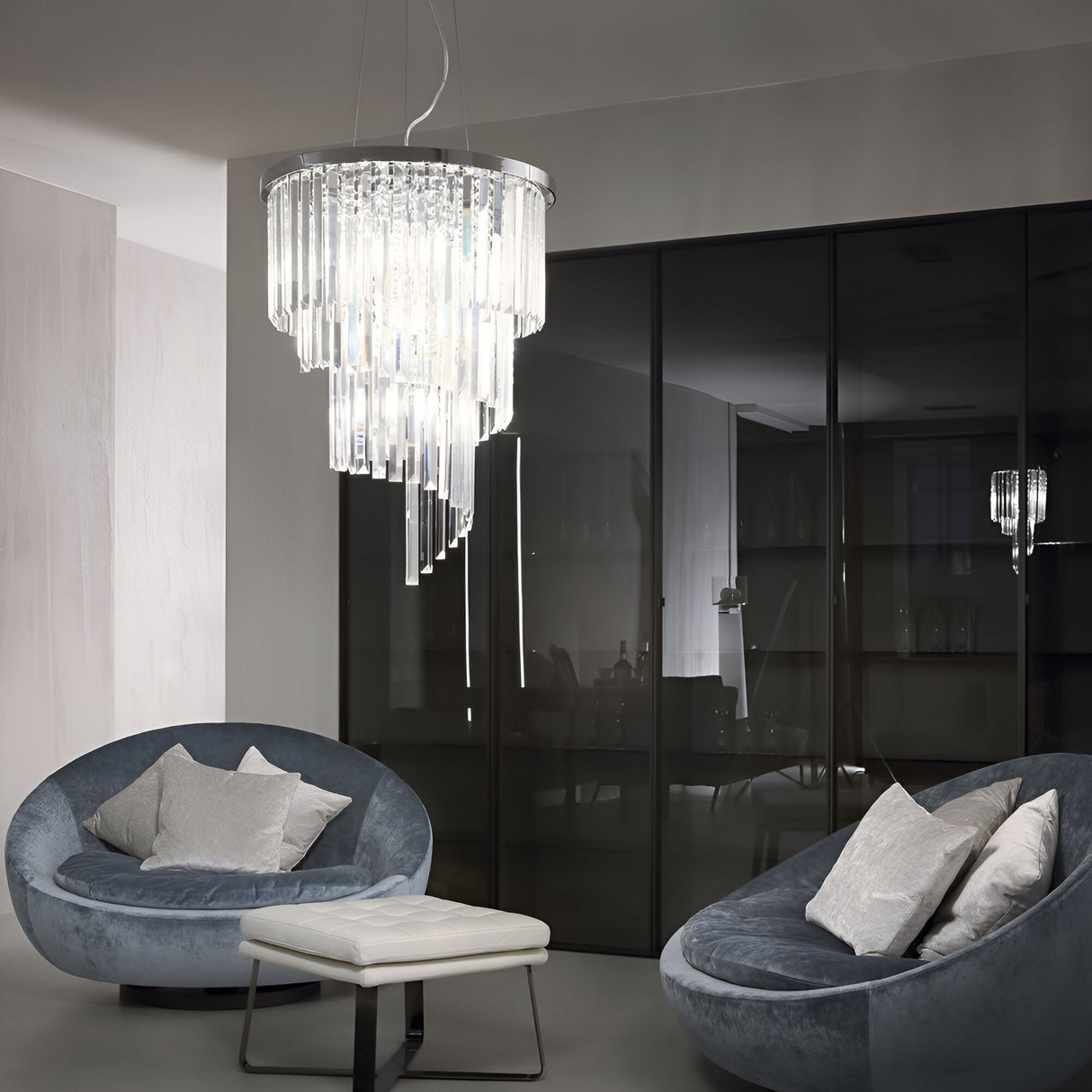 A modern living room showcases two round velvet chairs and a white cushioned stool under the Emberara Crystal 12 Light Chandelier in Chrome. Large glass panels in the background reflect the crystal lighting, enhancing an elegant ambiance with its dramatic fixture.