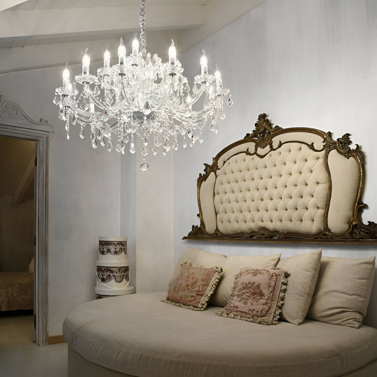 A luxurious bedroom showcases a round upholstered bed, floral-patterned pillows, and an ornate tufted headboard. The Grove Crystal 18 Light Chandelier in Chrome hangs above, casting a soft glow that enhances the elegance of the white-painted walls and decor.