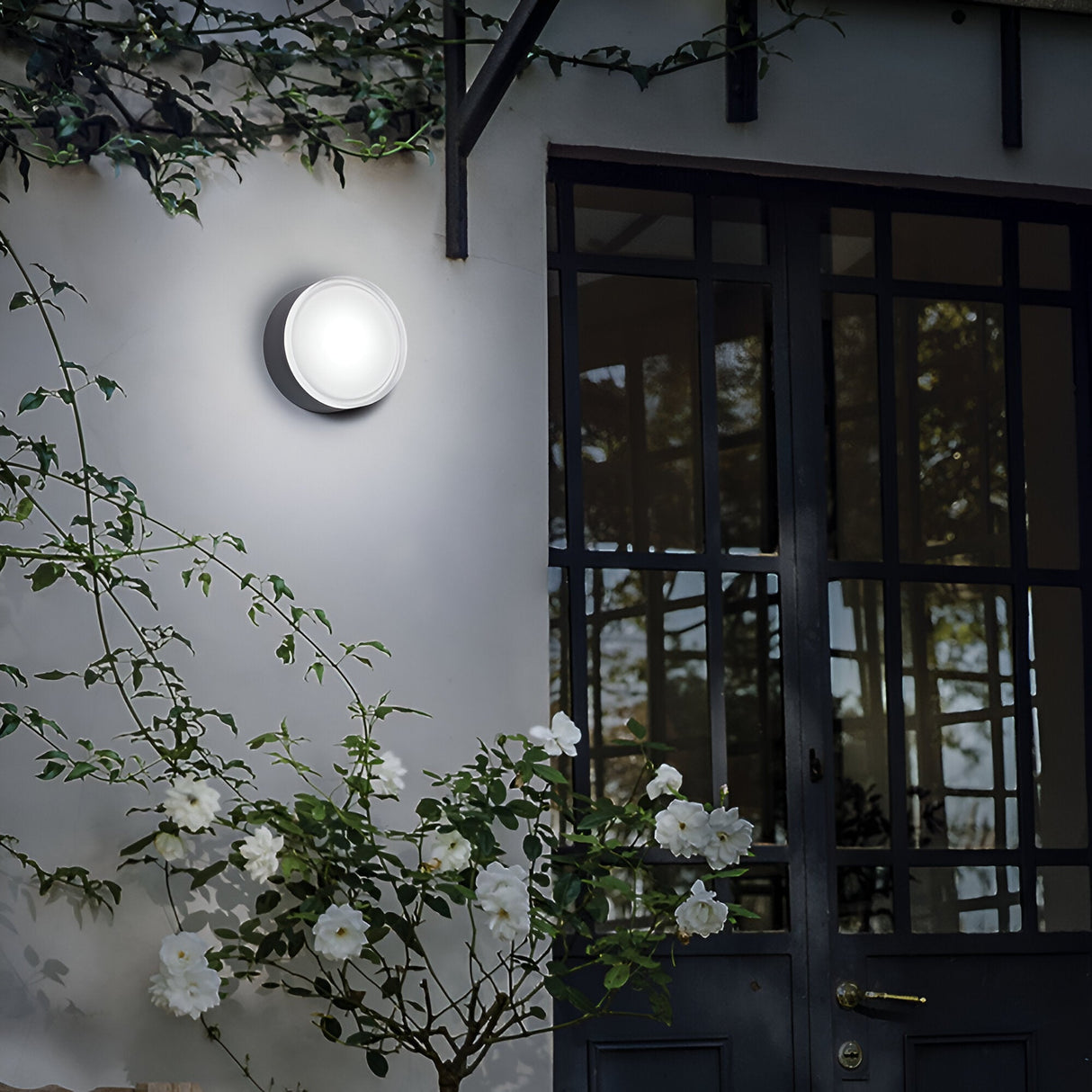 An Aura Small Flush Wall & Ceiling Light in white is illuminated on a wall next to a glass door. White roses with green leaves accent the foreground, while a metal trellis with climbing greenery frames the serene scene.