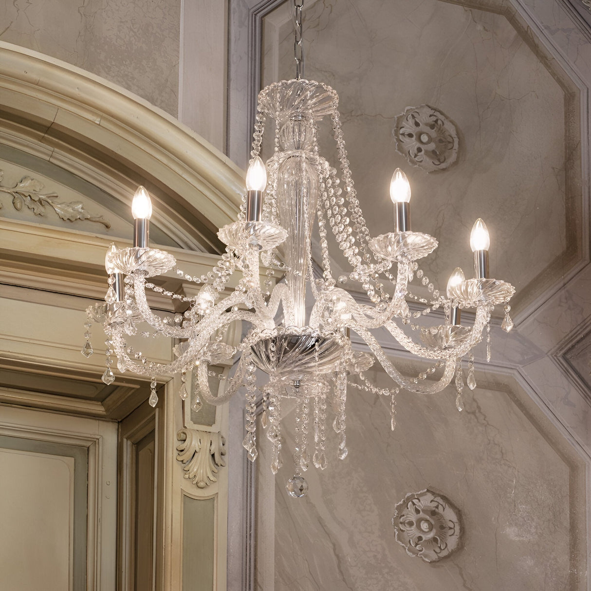 The Breeze-2 Crystal 8 Light Chandelier - Chrome, with its modern design and chrome finish, hangs from a ceiling adorned with ornate molding. This elegant chandelier boasts cascading crystal strands and intricate details, enhancing the room's classic decor with a touch of luxury.