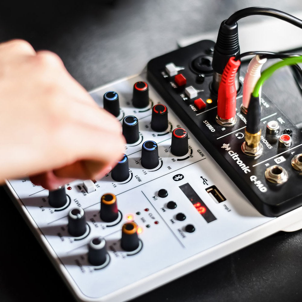 Hand adjusting the blue-ringed EQ knobs on the Citronic Q-PAD compact mixer by AVSL, with red and green XLR and jack cables connected at the rear.